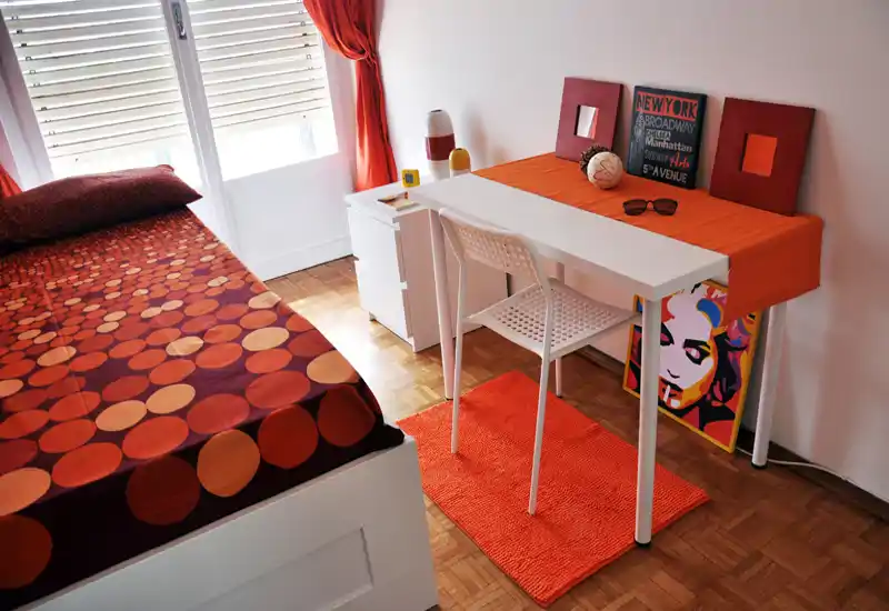 Bright, stylish bedroom with a single bed, white desk and warm orange accents including curtains, rug and bedcover; natural light from the window creates an inviting atmosphere.