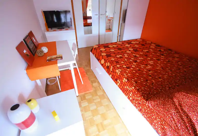 Bright, compact bedroom with a patterned orange bedspread, white storage bed frame and coordinated desk area; warm, well-composed decor and parquet flooring.