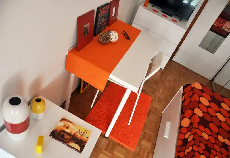 Bright, modern bedroom corner with a patterned orange duvet, small desk with orange runner and decorative accents — a cozy, well-styled small double or single room.