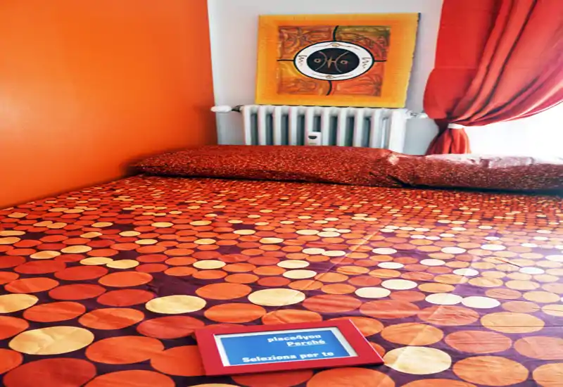 Bright, warmly styled bedroom with an orange-themed bedspread, matching curtains and decorative artwork above the radiator. The bed fills most of the frame, highlighting the cozy, well-composed sleeping area.