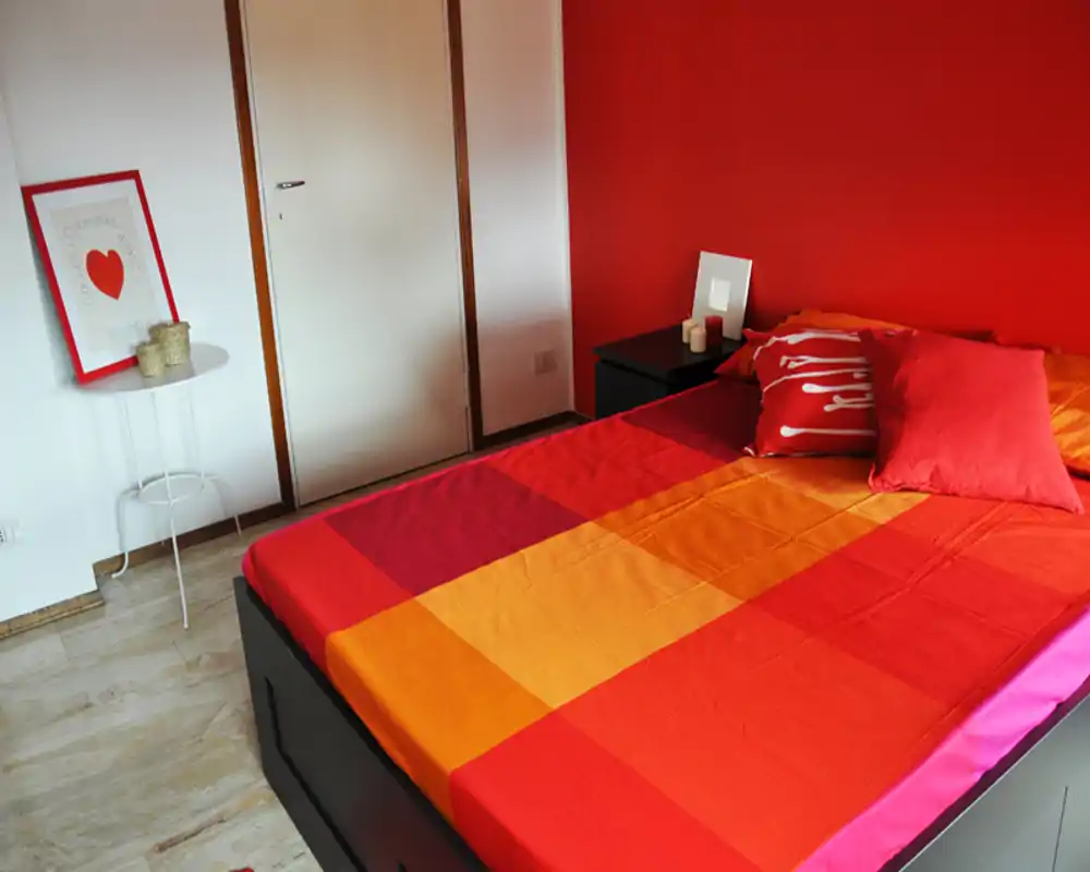 Camera da letto moderna e luminosa con un ampio letto colorato che occupa la maggior parte dell’inquadratura, parete rossa e comodino ordinato — perfetta per annunci contemporanei.