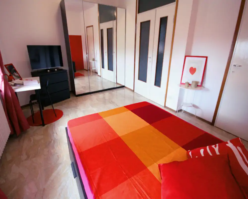Camera da letto luminosa e colorata con ampio copriletto rosso-arancione, armadio con ante a specchio, piccolo scrittoio e TV — accogliente e ideale per annunci.