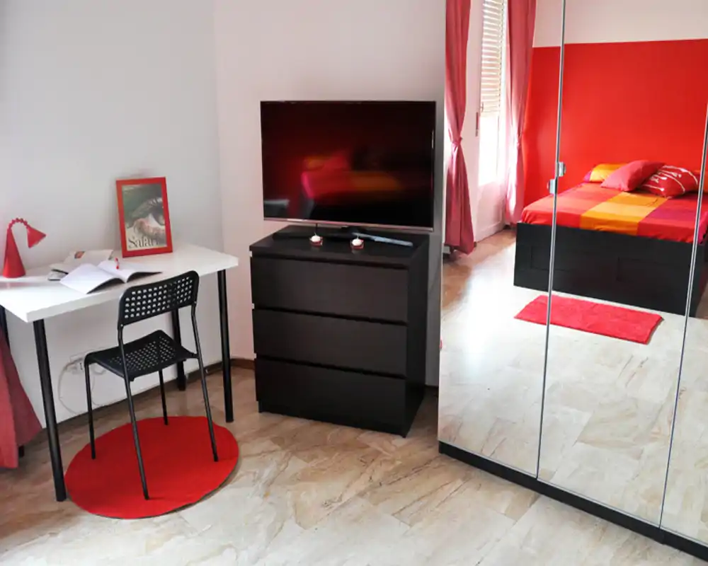Camera da letto luminosa e moderna con letto dai toni rossi, tessili coordinati e una cabina armadio specchiata che riflette parte della stanza. Comprende una TV su comò nero e una piccola scrivania vicino alla finestra.