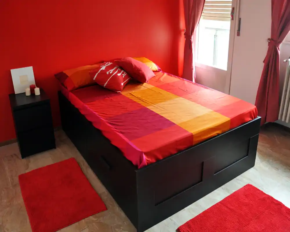 Camera da letto luminosa e colorata con un letto su pedana nero, biancheria rossa e arancione coordinata, tappeti abbinati e luce naturale da una finestra — un ambiente accogliente e ben arredato.