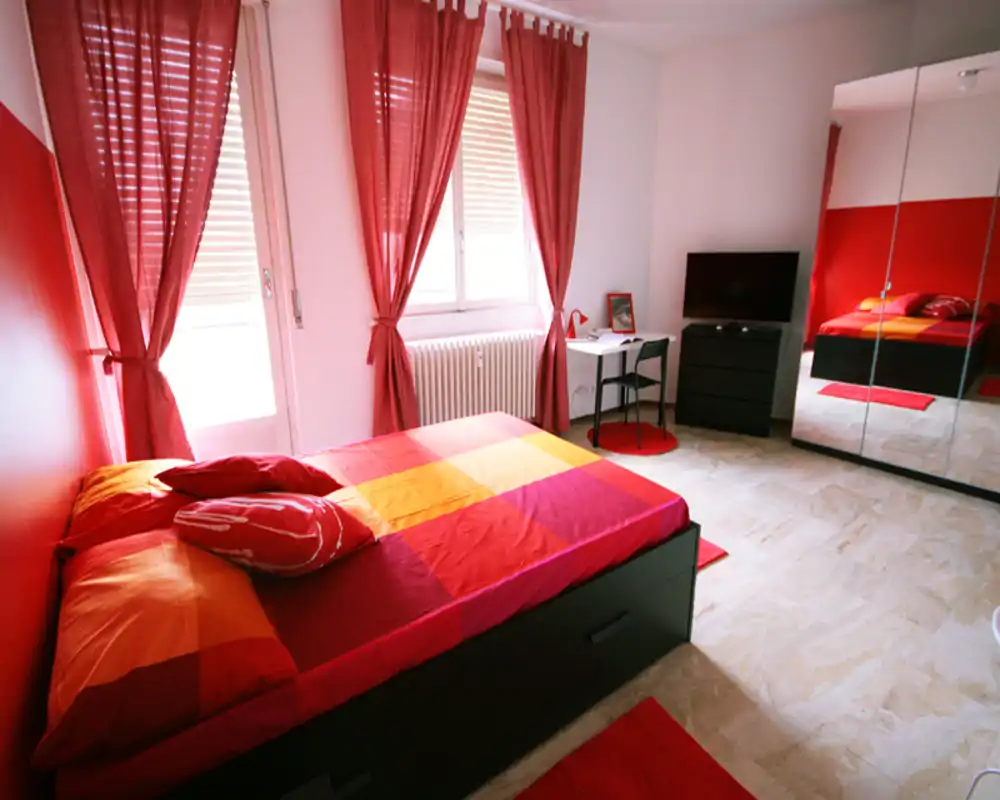 Camera luminosa e moderna con letto matrimoniale con biancheria rossa e arancione, ampia finestra con tende rosse, armadio con ante a specchio e una piccola scrivania — accogliente e di stile.