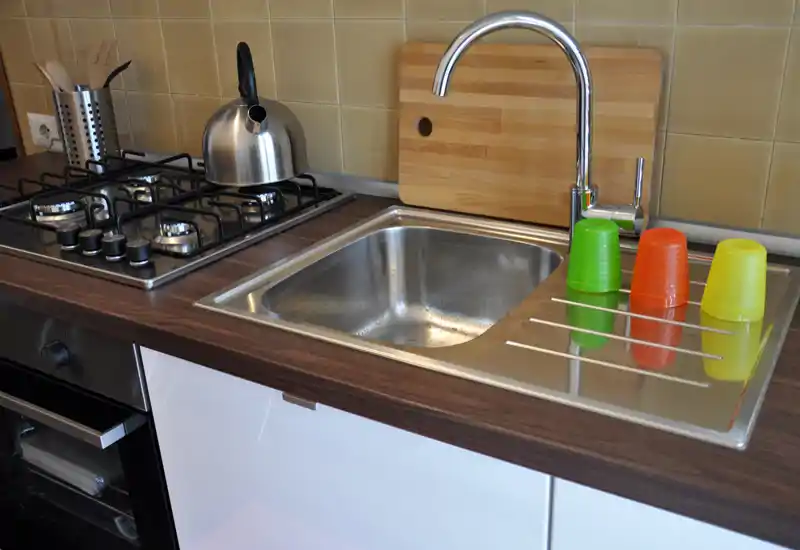 Cucina moderna compatta con lavello in acciaio inox, piano cottura a gas e piano in legno; bicchieri colorati e tagliere donano un tocco caldo.
