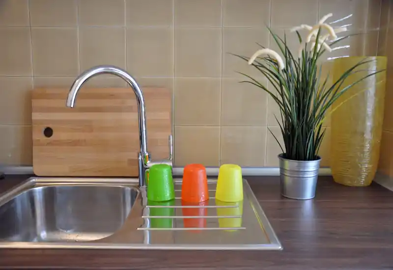 Zona lavello cucina luminosa e moderna con vasca in acciaio, rubinetto cromato, tagliere in legno, pianta decorativa e tre bicchieri colorati sull'asciugatoio.