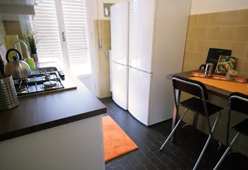 Cucina compatta e ben illuminata con piano cottura a gas, frigorifero bianco, piani scuri e un tappeto arancione; piccolo bancone colazione con due sgabelli.