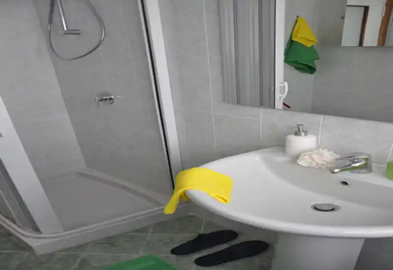Bagno compatto e pulito con doccia ad angolo, lavabo a colonna, specchio e piccoli accessori (dispenser, asciugamano, pantofole). Perfetto per un bagno funzionale in affitto.