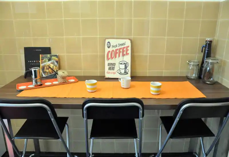 Bancone cucina compatto con tre sgabelli, piano in legno contro piastrelle, insegna decorativa del caffè e tazze su una runner arancione — un angolo colazione accogliente.