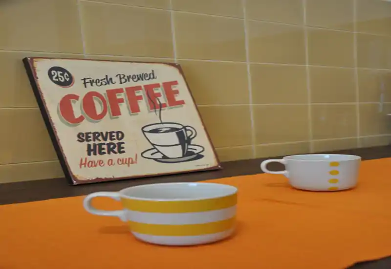 Primo piano di un angolo caffè: due tazze con motivo su un runner arancione e una vecchia insegna “Coffee served here” appoggiata su una piastrellata.