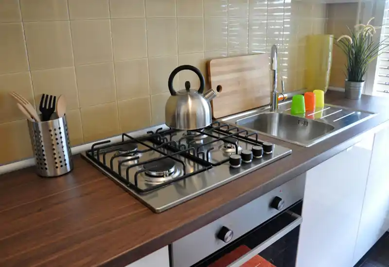 Angolo cucina moderno e luminoso con piano cottura a gas, lavello in acciaio e piano di lavoro in legno; accessoriato con bollitore, tagliere e dettagli decorativi.