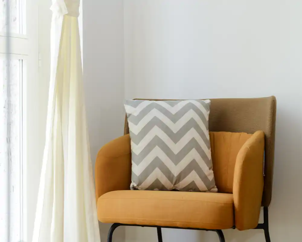 Fauteuil orange chaleureux près d'une fenêtre lumineuse avec un coussin à motif, parfait pour un coin lecture détente.