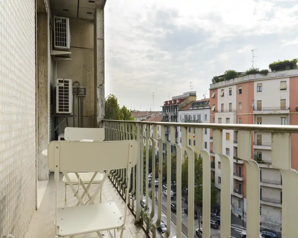 Balcon urbain ensoleillé avec petite table pliante et chaises, donnant sur une rue et des immeubles voisins — idéal pour un café le matin.