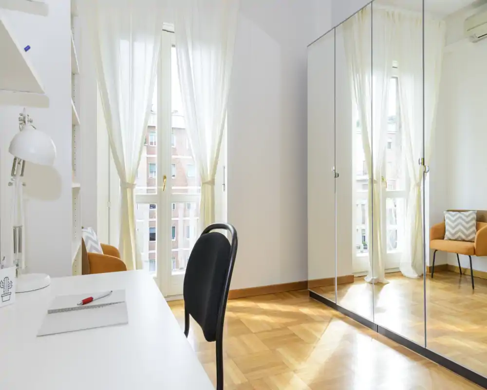 Coin de chambre lumineux et minimaliste avec grandes fenêtres, voilages et une armoire à portes miroir qui agrandit l'espace. Parquet chaleureux et un fauteuil d'appoint pour une atmosphère accueillante.