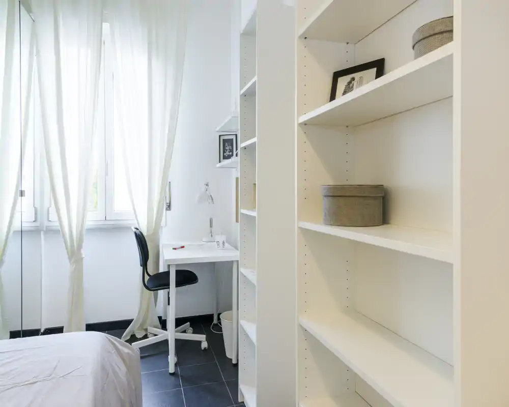 Bright, minimalist bedroom corner with a small desk and shelving. Natural light from large windows, neutral décor and simple bedding create a calm workspace within the sleeping area.