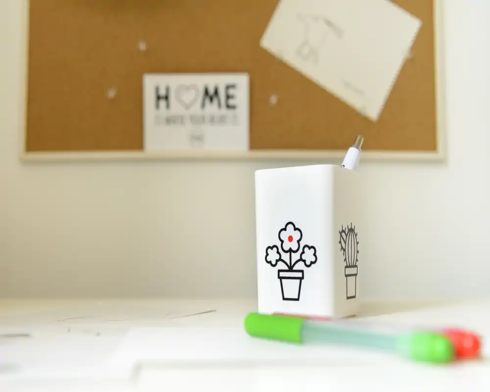 Close-up of a desk or workspace surface featuring a small white pen holder with simple plant icons and a few colored pens; a cork pinboard is visible blurred in the background.