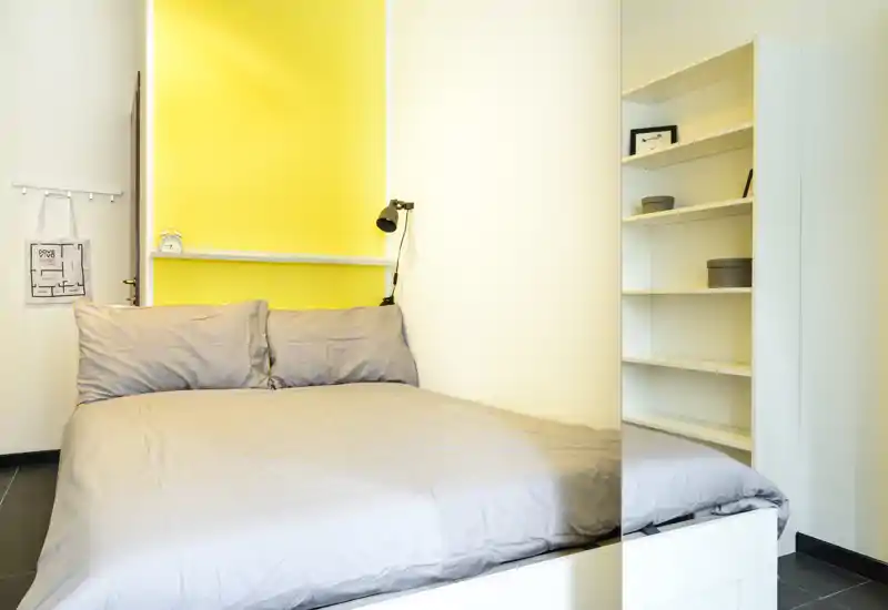 Bright, modern bedroom with a comfortable double bed, neutral bedding and a warm yellow accent wall. Built-in shelving offers practical storage and the lamp provides focused reading light.