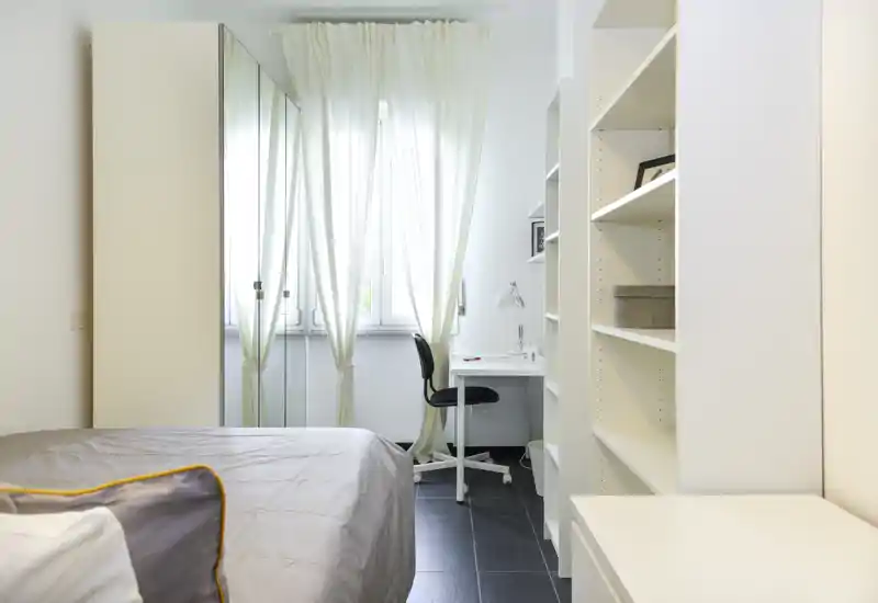 Bright, minimalist bedroom with a visible double bed in the foreground, sheer curtains over a large window and a small desk by the window — ideal for a compact, airy rental.