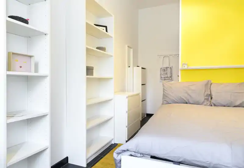 Bright, modern bedroom with a large bed, gray bedding and a cheerful yellow accent wall; built-in white shelving and drawers provide ample storage and a tidy look.