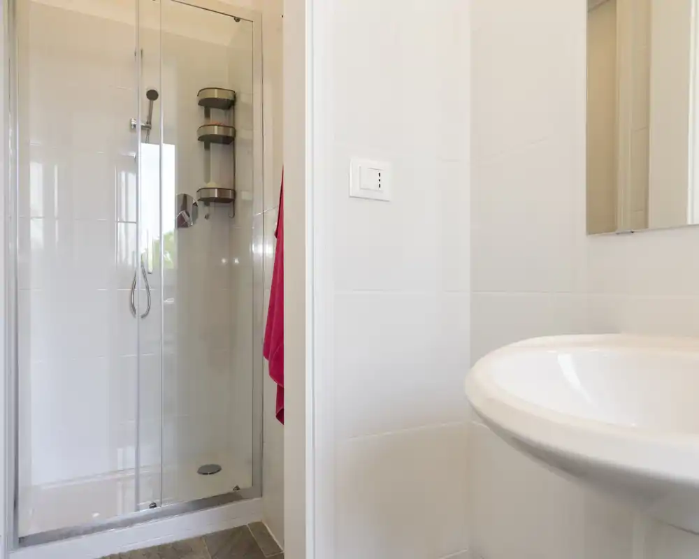 Clean, compact bathroom with a glass-enclosed shower and a pedestal sink; bright white tiles and a small mirror create a fresh, tidy feel.