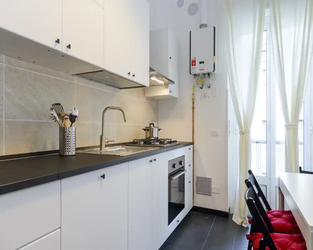 Modern fitted kitchen with a black countertop, stainless sink and built-in oven and gas hob. Natural light floods the room from a tall French-door window; bright tiles and simple white cabinets create a clean, functional space.