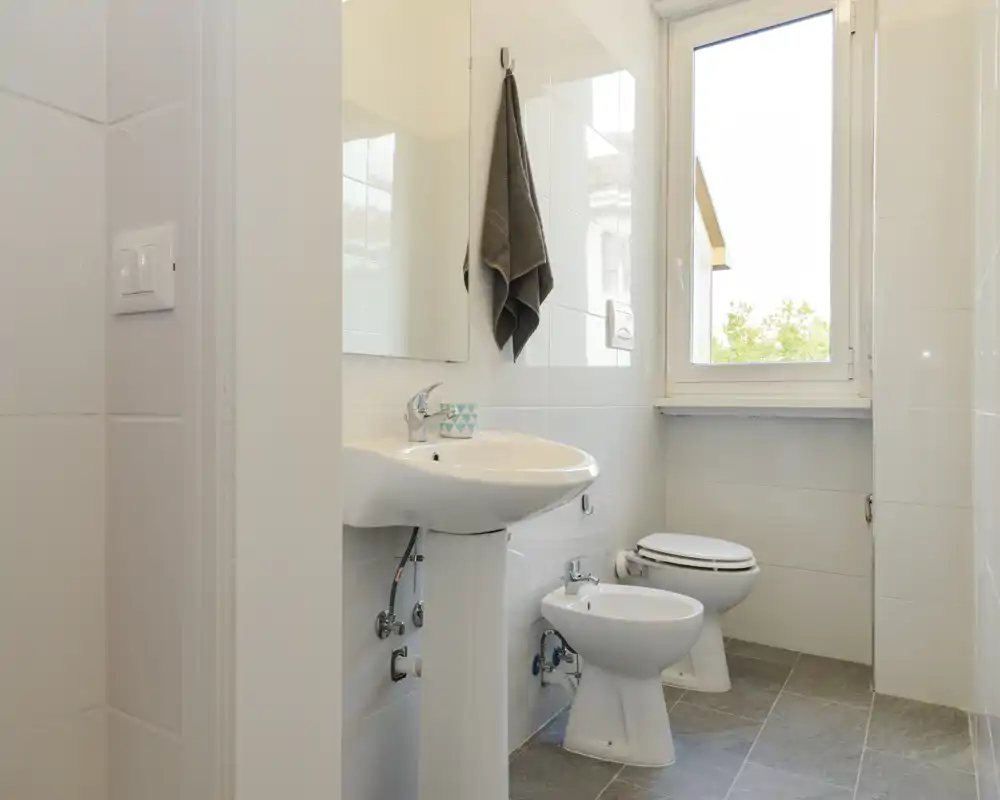 Compact, bright bathroom with a pedestal sink, bidet and toilet positioned under a large window; white tiles and natural light create a clean, airy feel.