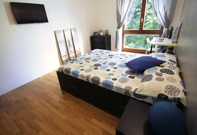 Bright, modern bedroom with a patterned double bed, wooden floors and a window letting in natural light. Simple desk and storage complete the practical, cozy setup.