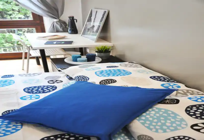Bright, well-styled bedroom corner with a patterned duvet and a decorative blue cushion; a small desk by the window offers a practical workspace and natural light.