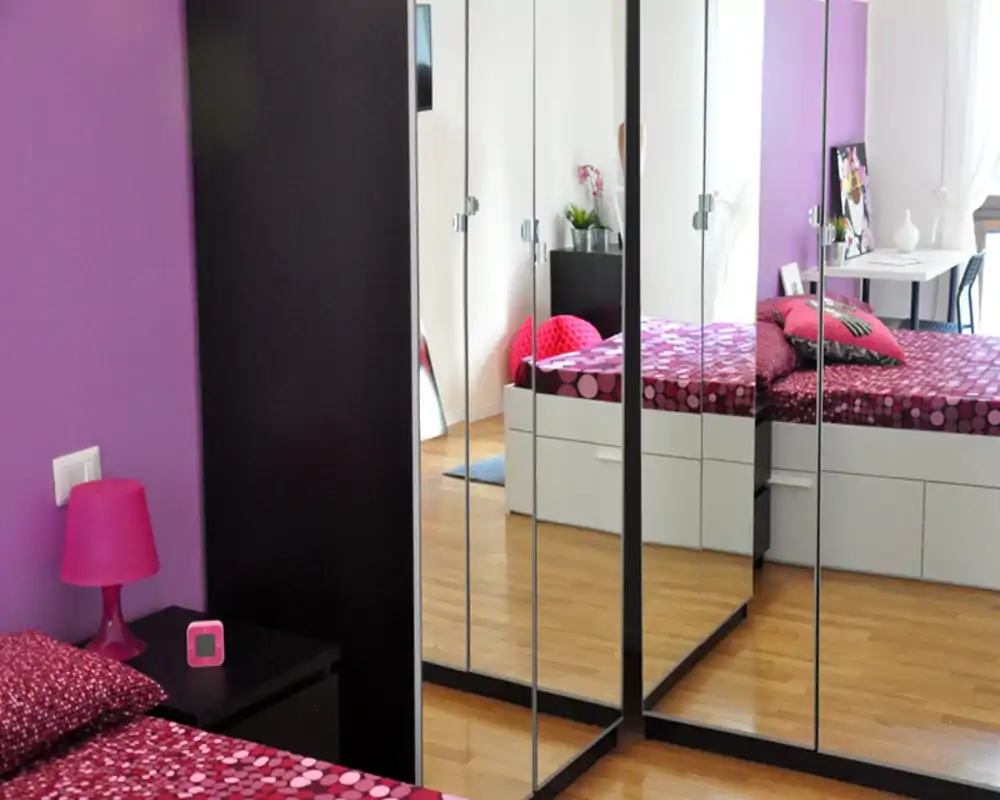 Camera da letto moderna e luminosa con copriletto a motivo rosa, cuscini decorativi e ante a specchio che ampliano visivamente l'ambiente.