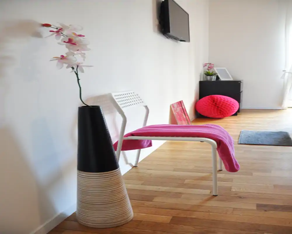 Ingresso/corridoio luminoso e minimal con parete bianca, pavimento in legno, una sedia con plaid rosa, un vaso decorativo e TV a muro — primo impatto ordinato e accogliente.