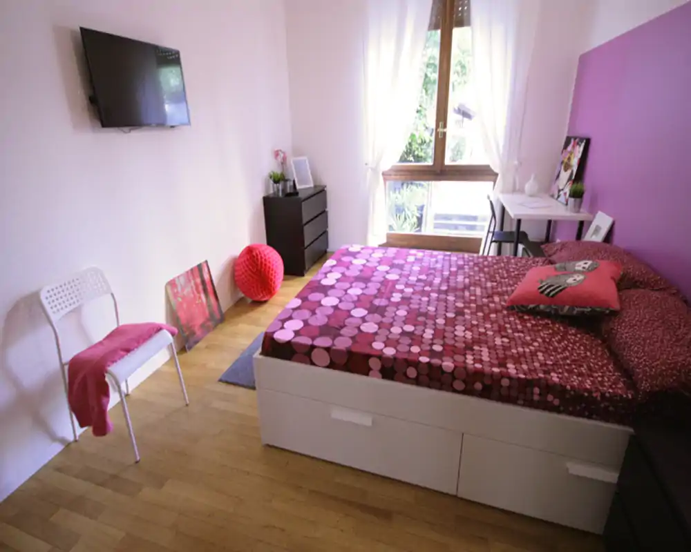 Camera da letto luminosa e curata con letto matrimoniale con copriletto magenta a motivo, una scrivania vicino alla finestra e pavimento in legno. La grande finestra offre luce naturale e un'atmosfera accogliente.