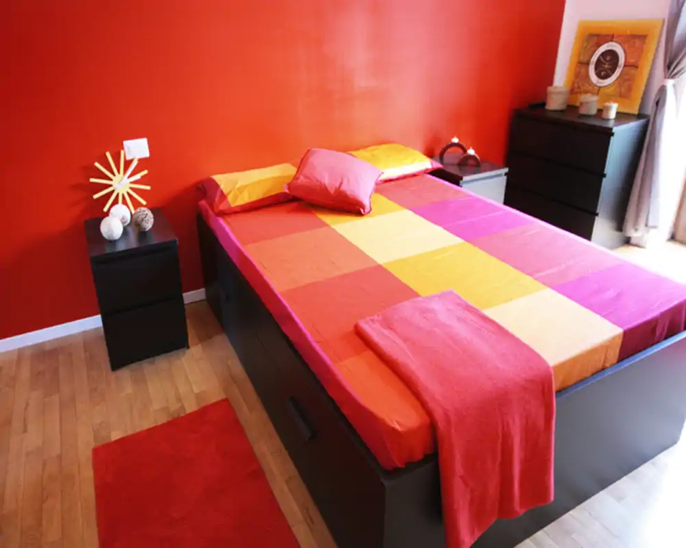 Dormitorio luminoso y moderno con una amplia cama doble vestida con ropa de cama roja, naranja y amarilla, alfombra a juego y mesitas de noche — ambiente acogedor y contemporáneo.