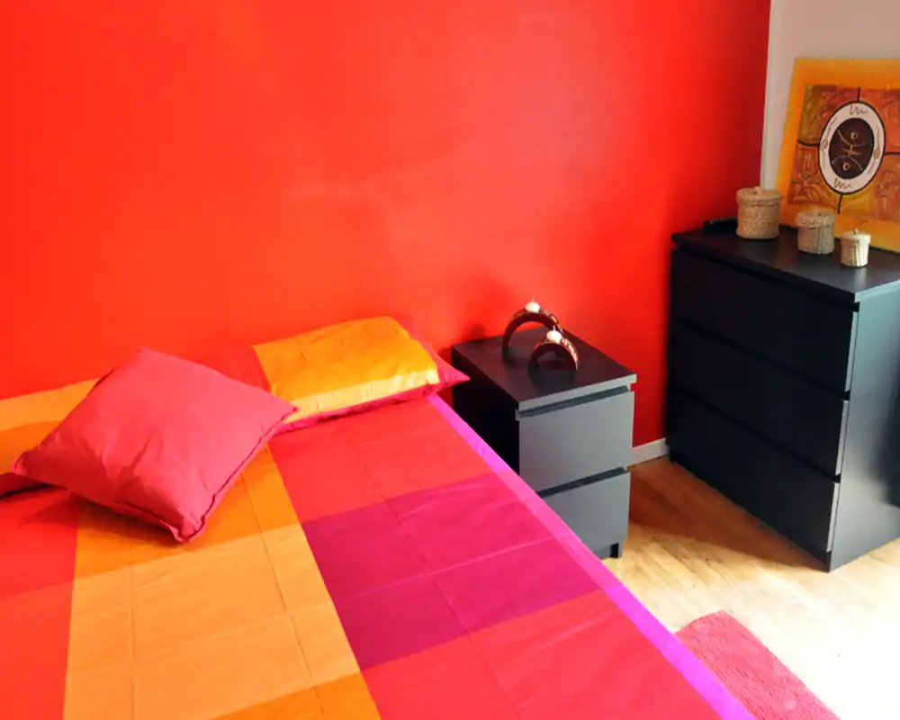 Dormitorio colorido con una gran cama vestida con ropa de cama naranja, rosa y roja, mesitas y cómoda en madera oscura. Espacio luminoso, cálido y bien presentado, perfecto para un anuncio de alquiler.