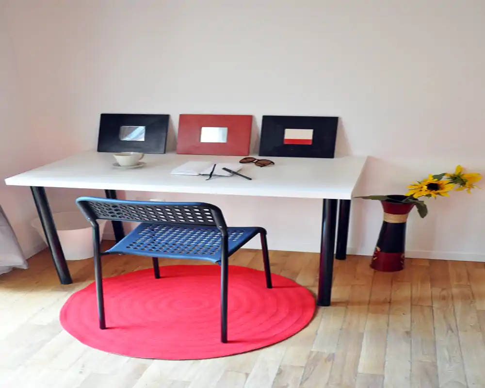 Espacio de trabajo compacto y luminoso con escritorio blanco, silla perforada y alfombra circular roja; espejos decorativos y un jarrón con girasoles aportan carácter.