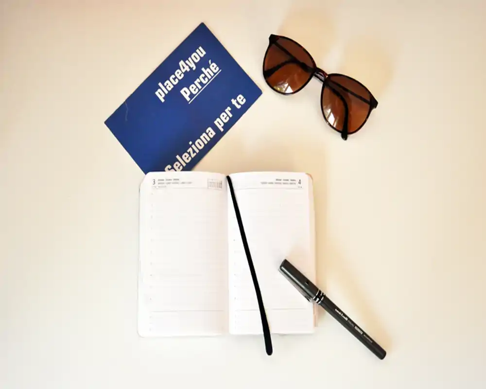Plano cenital de un cuaderno abierto, un bolígrafo, gafas de sol y una tarjeta azul sobre una superficie clara — una composición de escritorio.