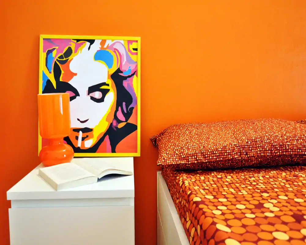 Angolo di camera luminoso e moderno con pareti arancioni vivaci e biancheria a pois; il comodino ospita una lampada, un libro aperto e una stampa pop-art colorata.