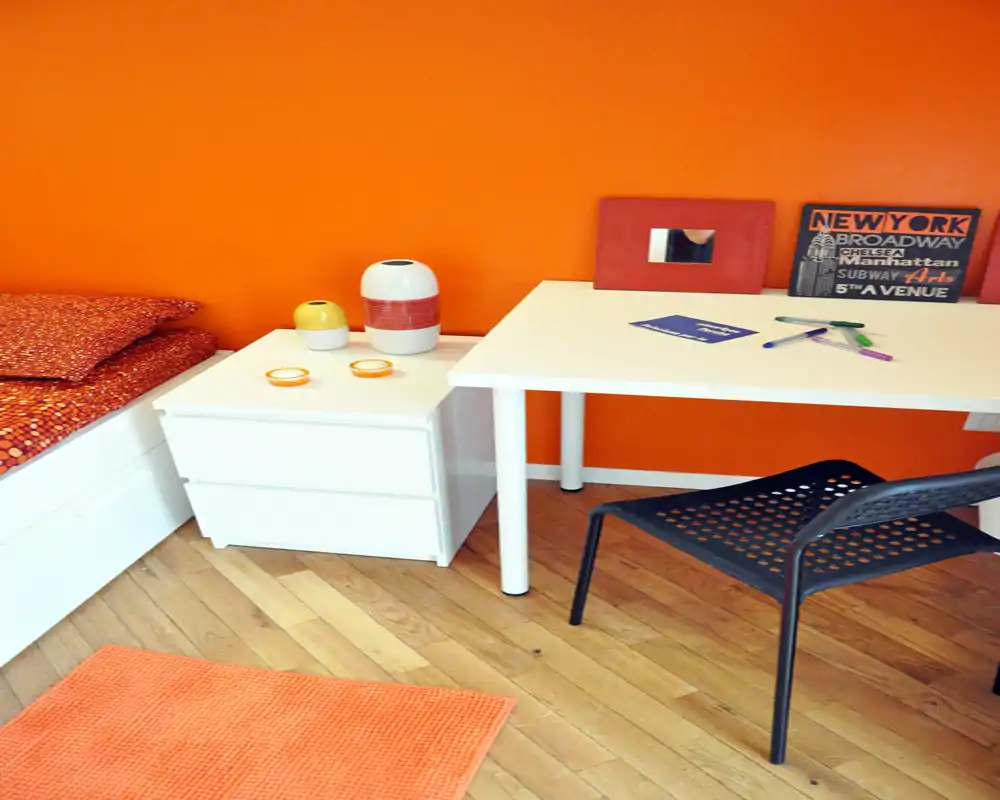 Angolo di camera da letto luminoso e moderno con scrivania bianca, cassettiera e parete arancione; biancheria in ordine e oggetti decorativi creano un'atmosfera calda e contemporanea.