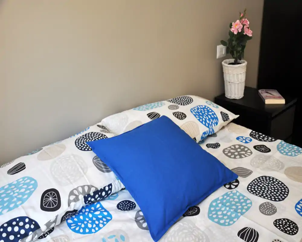 Rincón de dormitorio luminoso y ordenado con ropa de cama estampada y un cojín decorativo azul sobre la cama junto a una mesita con planta.