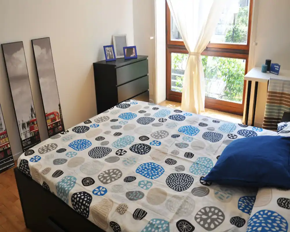 Dormitorio luminoso y bien decorado con ropa de cama estampada, una cómoda negra baja y una gran ventana que aporta luz natural — limpio y acogedor.