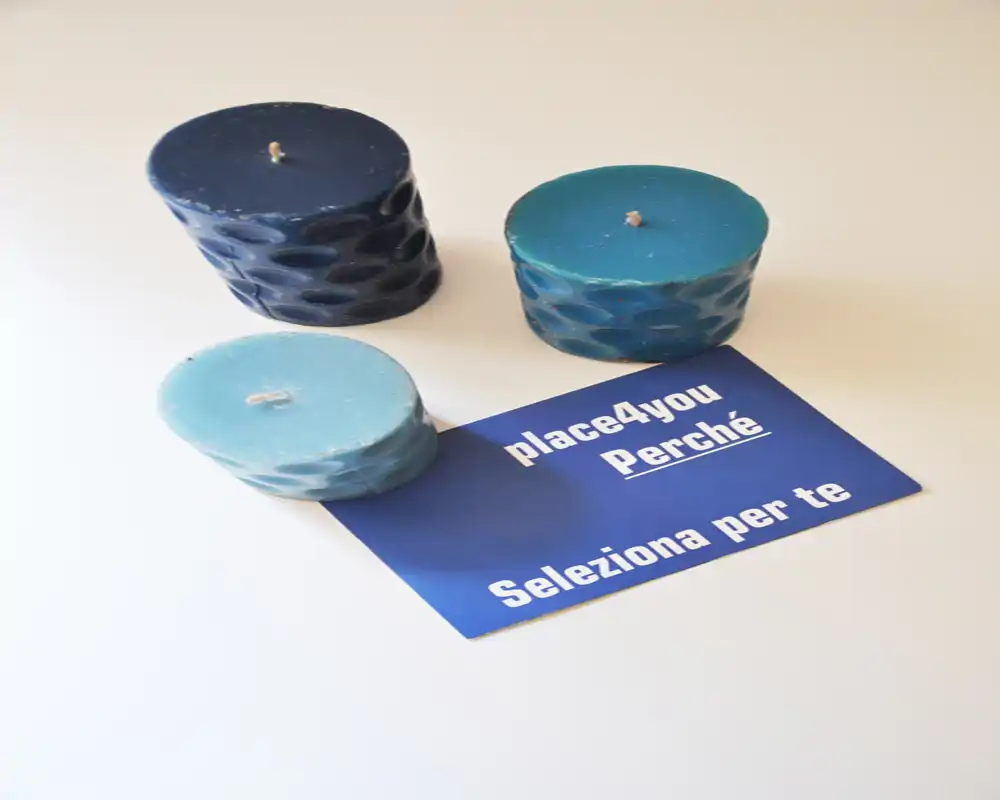 Primer plano de tres velas decorativas azules sobre una superficie clara junto a una tarjeta azul que dice 'place4you Perché Seleziona per te'. La composición enfatiza las velas como objetos decorativos sobre una mesa.