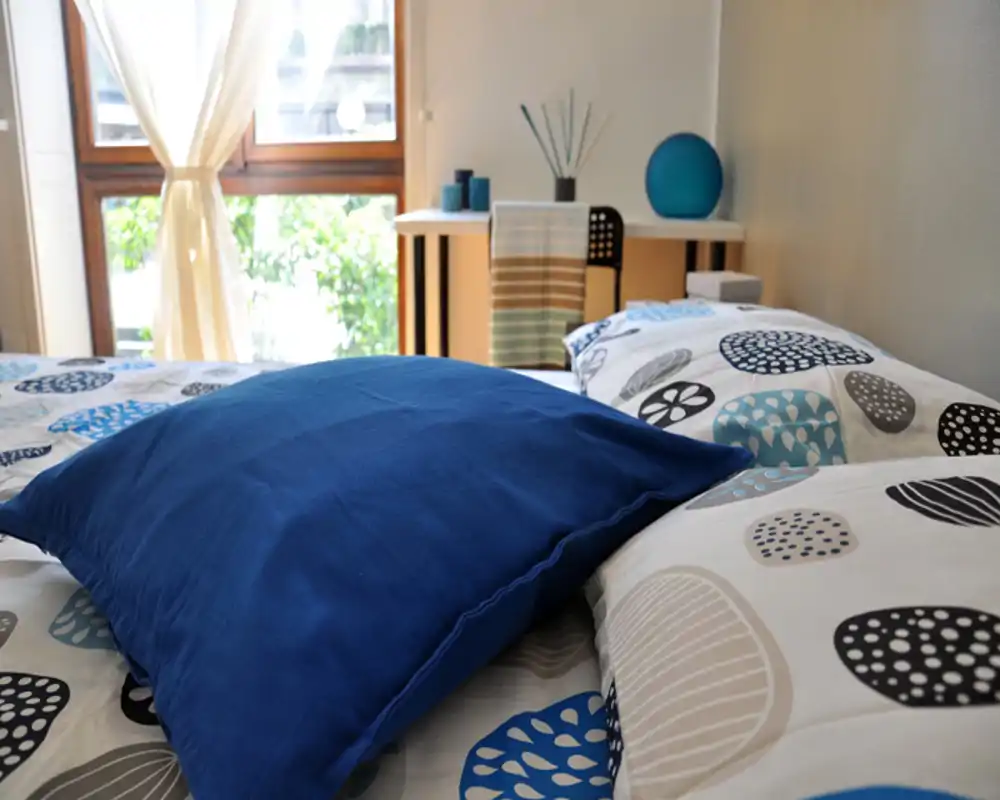 Dormitorio luminoso y acogedor centrado en la ropa de cama estampada y un gran cojín azul en primer plano, con luz natural desde la ventana y una pequeña mesa en el fondo.