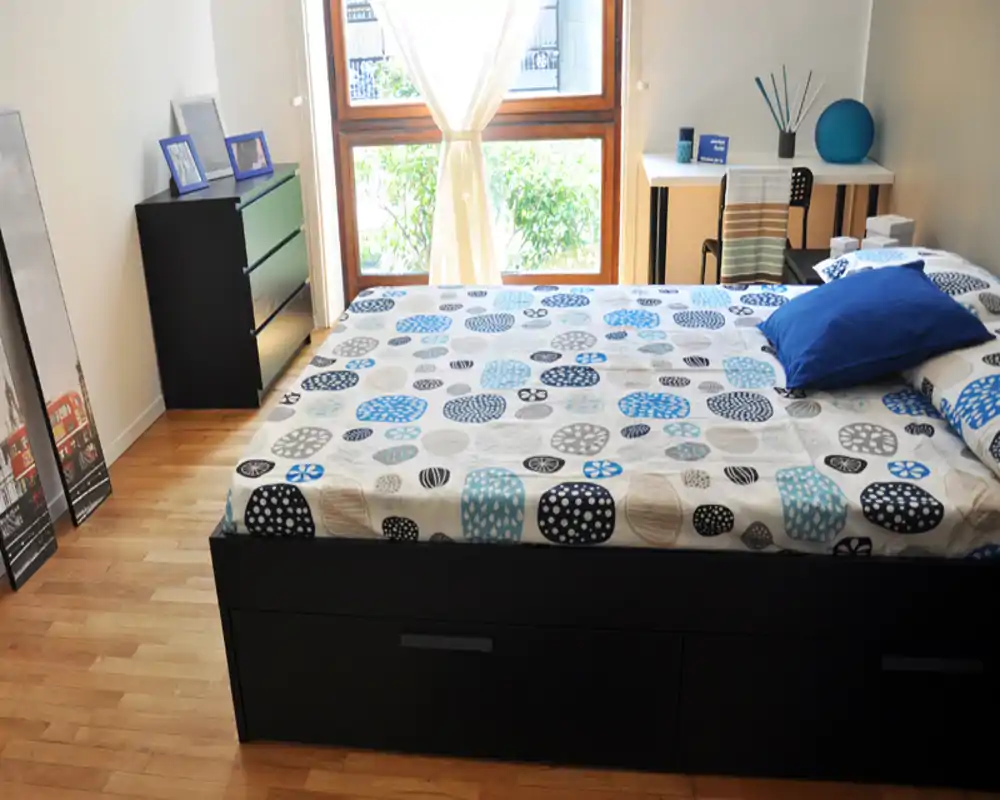 Dormitorio moderno y luminoso con una cama doble grande, edredón estampado y luz natural procedente de la ventana. Mobiliario sencillo y ordenado con cómoda y pequeño escritorio.