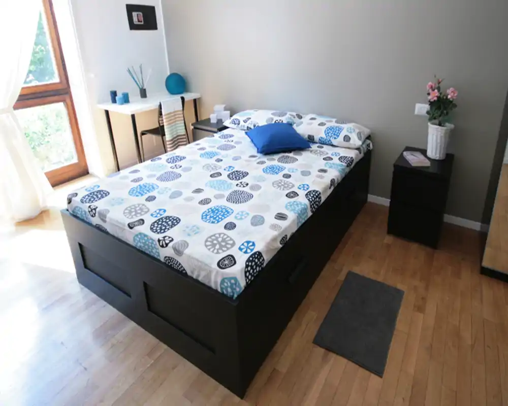 Dormitorio moderno y luminoso con una cama doble grande, ropa de cama estampada, mesita de noche y luz natural de una ventana alta. Suelo de madera limpio y sutiles detalles decorativos que crean un ambiente acogedor.