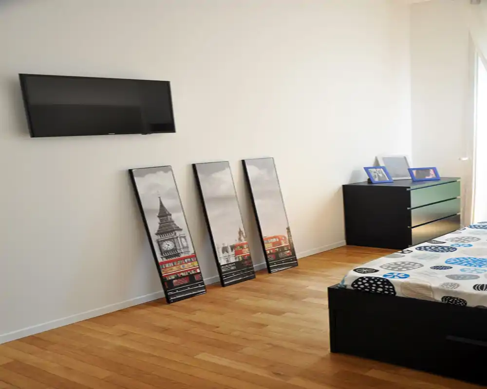 Dormitorio moderno y luminoso con edredón estampado, estructura de cama baja negra y cómoda, televisor en la pared y cuadros apoyados contra la pared — espacio de descanso limpio y minimalista con luz natural.