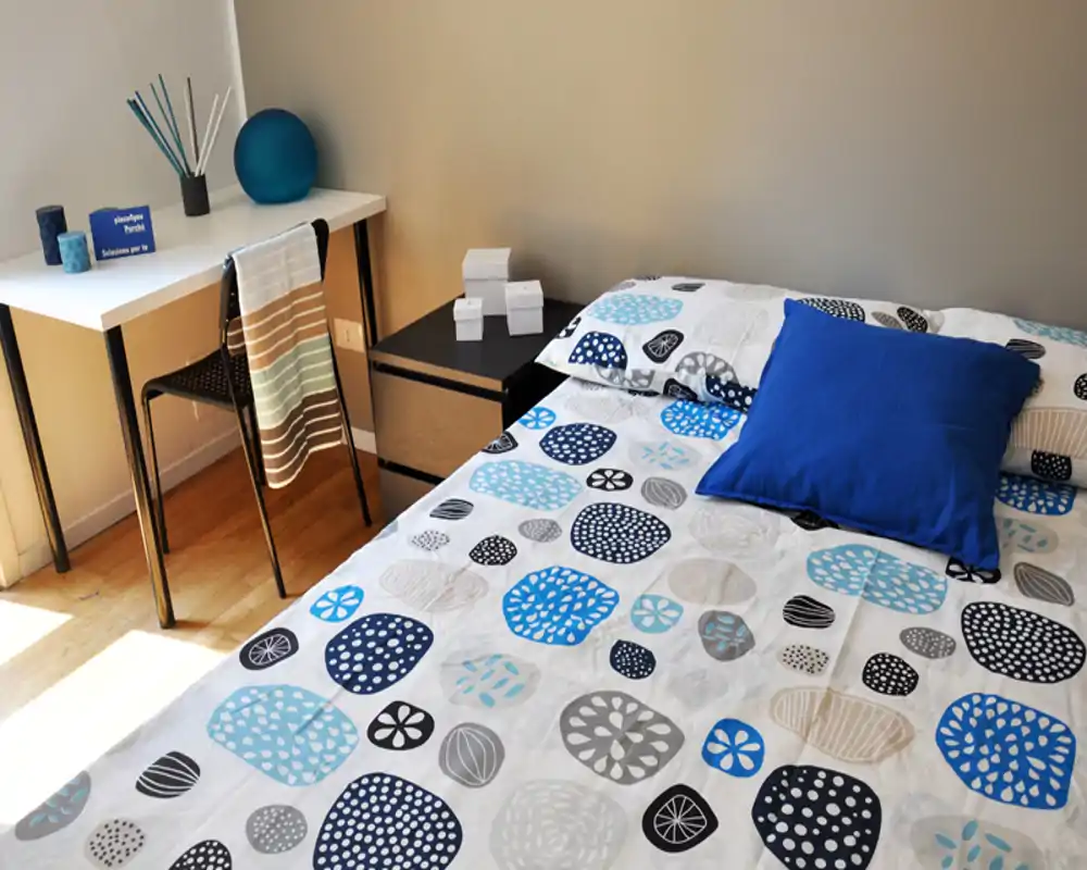 Dormitorio luminoso y ordenado con edredón estampado y cojín azul; un pequeño escritorio y silla junto a la ventana ofrecen un espacio de trabajo práctico.