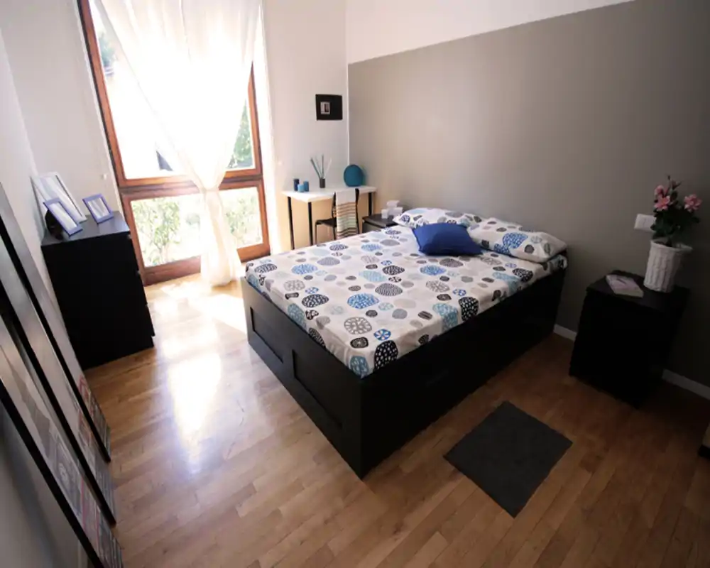 Dormitorio doble luminoso y bien decorado con edredón estampado, mesillas y una gran ventana que aporta abundante luz natural — perfecto para anuncios de alquiler.