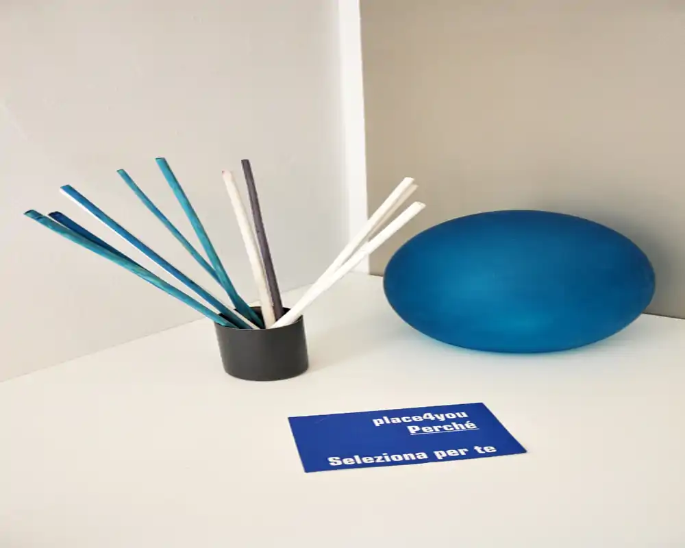 Primer plano de decoración sobre una superficie: palitos difusores en un pequeño soporte, un adorno esférico azul y una tarjeta pequeña sobre una superficie blanca; la foto se centra en accesorios, no en la habitación.