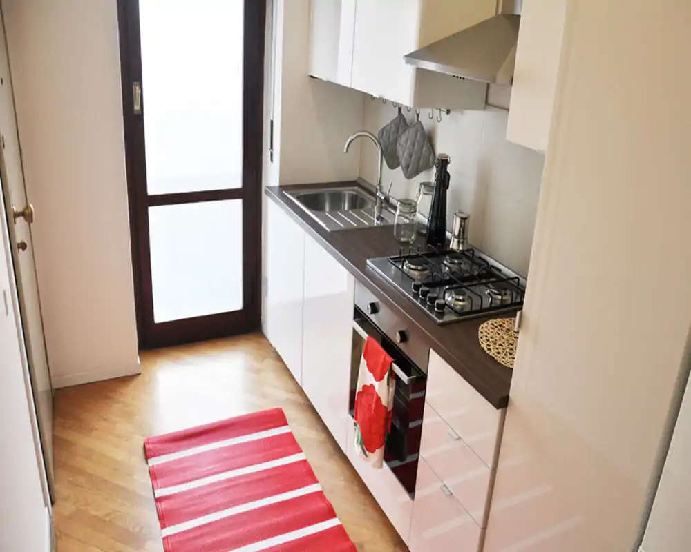 Cocina moderna y compacta con muebles blancos lacados, encimera oscura, placa de gas de cuatro fuegos y fregadero de acero; luz natural y detalles rojos que aportan calidez y funcionalidad.