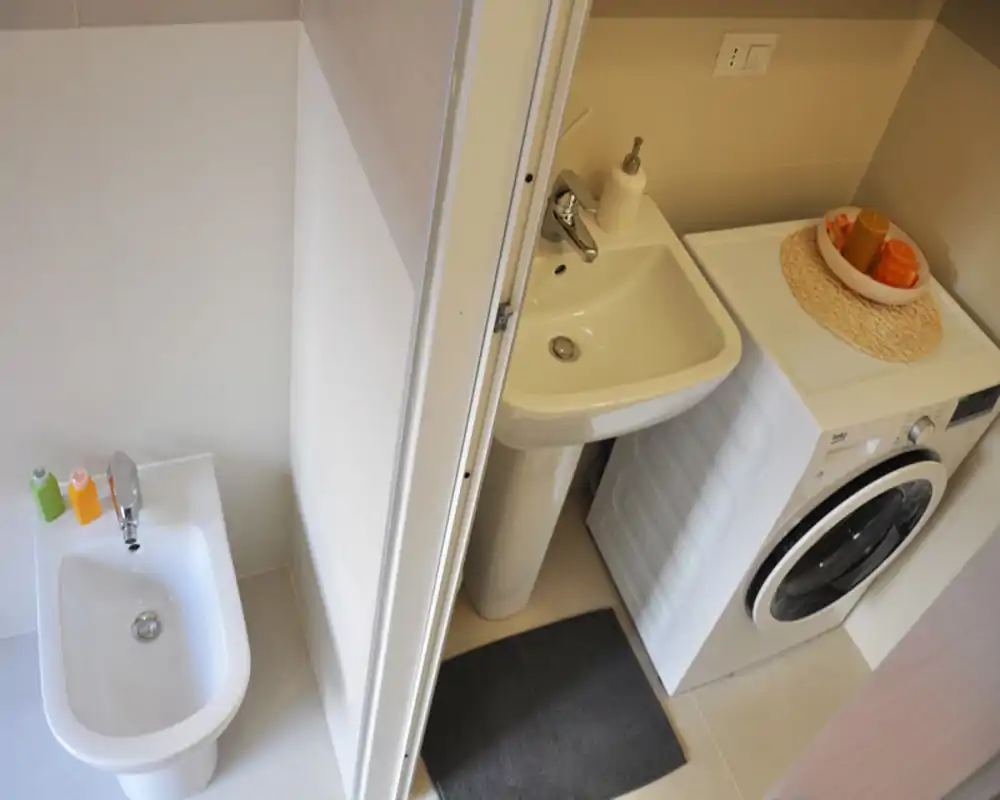 Rincón de baño moderno y compacto con bidé, lavabo con pedestal y lavadora de carga frontal visto desde arriba. Baldosas neutras y accesorios sencillos ofrecen una sensación ordenada y funcional.